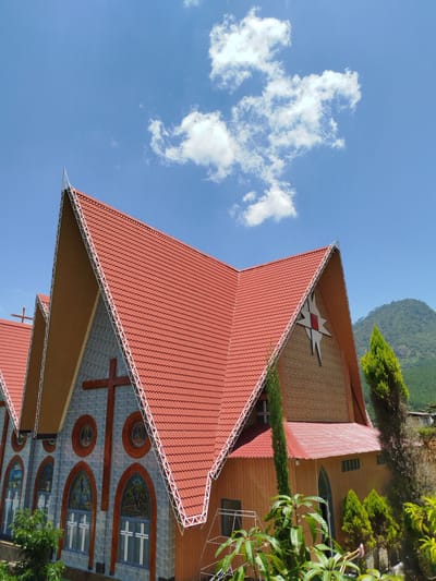 The image shows a church with a red tiled roof and a large cross on its facade. The church is surrounded by lush green trees and a mountain range in the background. The sky is blue with some scattered clouds. The church appears to be made of stone and wood. The architectural style is somewhat gothic with pointed arch windows. A star-shaped emblem is visible on the upper part of the church. The scene suggests a peaceful and serene environment. The specific location in K'Rèn, Vietnam, is not explicitly shown but the mountainous landscape and vegetation are consistent with such a region. There are no people visible in the image.