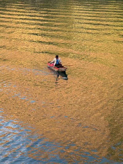 A single individual navigates a small wooden boat on the waters of Lake Atitlán, near San Pablo La Laguna, Guatemala. The person is seated within a red-painted, simple wooden boat, holding a wooden oar or paddle in the right hand, positioned to propel the craft. They are dressed in a dark cap, a light-colored long-sleeved jacket over a blue shirt, and dark trousers. A pink object is visible inside the boat, near the bow. The water's surface reflects an intense golden-orange hue, likely from a low-angle sun or reflections of surrounding landscape, creating ripples across the majority of the visible area. Patches of blue-grey water are visible in the lower left, indicating areas not dominated by the golden reflection.