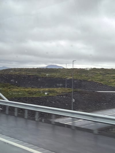 The image depicts a cloudy, overcast day in a rugged, natural landscape, likely on the outskirts of Hafnarfjordur, Iceland, near a roadway. In the foreground, a metal guardrail runs horizontally across the frame, separating a wet asphalt road with white lane markings from the elevated terrain behind it. The road surface appears slick, suggesting recent rain or damp conditions.

Beyond the road, the landscape rises sharply, characterized by dark, rocky formations interspersed with patches of green, moss-like vegetation. This layered topography creates a textured, uneven surface that extends into the midground. A thin, dark line, possibly a road or pathway, is visible running along one of the elevated ridges.

In the background, the terrain continues to roll, with more vegetation and dark soil visible. Distant mountains, appearing as muted blue shapes against the horizon, are partially obscured by the low-hanging clouds. The sky is completely covered by a thick layer of grey clouds, with no discernible sunlight. Several tall, slender streetlights, typical of Icelandic roads, are positioned along the ridgeline, some equipped with modern, angled lamp heads. One streetlight is particularly prominent in the mid-right of the frame. The overall impression is one of a stark, windswept environment under a typical Icelandic sky. No people or vehicles are visible in the frame.