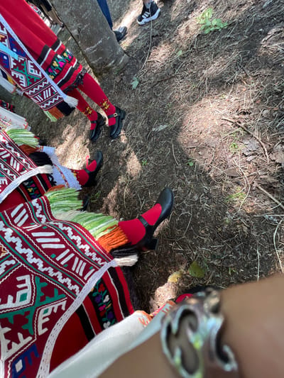 The image captures an outdoor scene in Bolyarovo, Bulgaria, observed from a low perspective. In the immediate foreground, the lower portion of a human arm or wrist, adorned with a prominent silver bracelet featuring an ornate design, is visible.
Two individuals are present, partially visible, dressed in traditional folk costumes. The figure on the left wears a long red garment, possibly a skirt or apron, embellished with horizontal bands of black fabric embroidered with colorful floral and geometric patterns, and trimmed with white lacework. Their legs are covered in red tights, with the ankles featuring distinct yellow and gold stylized leaf embroidery. They wear black Mary Jane-style shoes secured with gold buckles. A white woven strap or sash with multi-colored geometric patterns hangs diagonally from this costume.
The second figure, positioned on the right, is largely obscured, but their lower body displays a vibrant garment. This includes a dark red fabric with white and green geometric embroidery, accompanied by multi-colored fringes in shades of white, green, and orange yarn. One foot is visible, clad in a red tight and a black shoe, similar in style to the other individual's.
The ground surface consists of dry, exposed earth mixed with fallen leaves, small twigs, and scattered patches of green vegetation. Two tree trunks are centrally located in the mid-ground, indicating a wooded or natural environment. In the blurred background, other individuals are faintly visible, including one person wearing blue jeans and black sneakers with white soles. The scene is brightly lit by natural sunlight, casting shadows on the uneven ground.