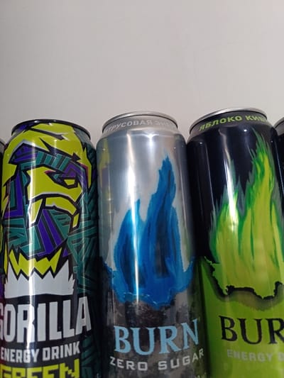 Three energy drink cans are displayed close together. The can on the left is yellow and green, featuring an image resembling a gorilla. The middle can is silver, with a blue flame illustration and "Burn Zero Sugar" text. The can on the right is dark, with a green flame design and "Burn Energy Drink" text. The tops of the cans are slightly visible, each with inscriptions in a language that appears to be Russian.