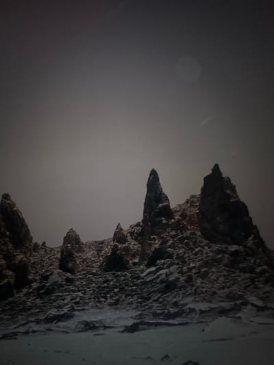 This is a vertical, low-light, close-up photograph of a jagged, rocky cliff face. The rocks are dark and textured, with sharp, irregular edges and protrusions. The cliff is positioned on the right side of the frame, taking up approximately the right half of the image. The left side of the image is a gradient of dark gray to a lighter, hazy gray.
In the upper left quadrant of the image, a faint, circular, lighter shape is visible against the dark background, possibly representing a moon or a distant light source through haze. The overall lighting is dim, suggesting a nighttime or very overcast setting. The texture of the rocks suggests a natural, possibly arid or eroded geological formation. The image's orientation is rotated, with the cliff face appearing to descend from the top of the frame towards the bottom. There is no text visible in the image. The scene appears to be an outdoor, natural environment.