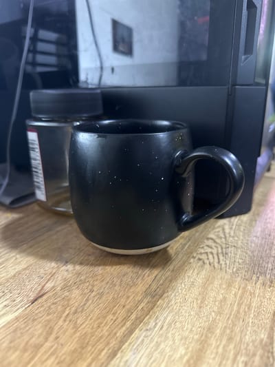 This is a close-up, rotated, eye-level shot of a black speckled mug resting on a light brown wooden surface. The mug has a curved handle and its interior appears to be lighter in color. Some white speckles are visible on the black exterior of the mug. Partially visible to the left of the mug is a black electronic device, possibly a printer or a computer tower, with a reflective surface. A barcode is faintly visible on a white label attached to a silver object beneath the mug. The wooden surface shows grain patterns and some slight discoloration or wear. The lighting appears to be artificial and casts subtle shadows. No people or animals are present in the image, and the scene is indoors. The overall mood is neutral, focusing on the objects and their immediate surroundings. No specific time of day or weather conditions are discernible. The location context "Juan Mina, Colombia" does not provide any visual cues within the image itself.