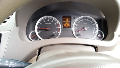 The image shows a close-up view of a car's dashboard, specifically the instrument cluster and steering wheel. The instrument cluster features a tachometer on the left, a digital display in the center, and a speedometer on the right. The digital display shows the gear (D), average fuel consumption (13.2 km/L), distance covered (100 km), and time (4:53). A small fuel gauge is visible to the right of the speedometer. The steering wheel, covered in a beige material, is partially visible at the bottom. The overall impression is of a car interior, likely parked. No people are visible. The image is a photograph taken from inside the car, looking towards the dashboard. There is no information within the image to indicate the location in the city of Blangrheuem, Indonesia.