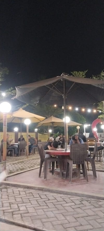 An outdoor seating area at night in Meunasahgeumata, Indonesia, appears to be a restaurant or cafe. Several tables and chairs are visible, some occupied by people. A large black umbrella shades a central table where two individuals are seated, one with their head down on the table. Other diners are visible at tables in the background, some engaged with their phones. String lights are hung across the scene, and large globe lights are present on poles. The ground is paved with interlocking bricks. The overall atmosphere suggests a casual evening gathering.