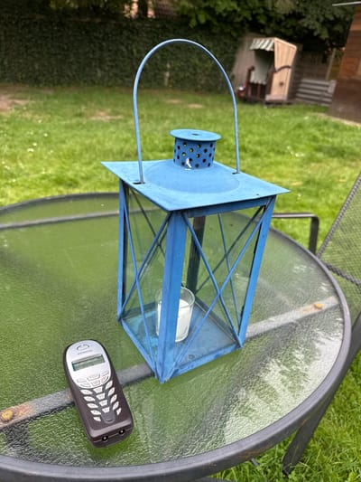 An overhead shot shows a blue metal lantern with a glass body resting on a round glass-top table. The lantern has a handle and a perforated cap at the top. Inside the lantern, a white candle is visible. A black Siemens mobile phone lies on the table next to the lantern. The table is positioned on a grassy lawn, with a hint of a wooden fence and a covered seating area visible in the background. The lighting suggests it is daytime, and the greenery indicates an outdoor setting, likely a garden. The scene appears to be in Wiesmoor, Germany.