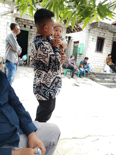 The scene depicts an outdoor gathering in Juwana, Indonesia. In the center, a man with short dark hair, wearing a black and white patterned batik long-sleeved shirt and black trousers, holds a baby dressed in a light-colored outfit and a beige cap. The man gazes at the baby, who is looking towards the viewer with a hand near its mouth.
To the left, another man in a light grey long-sleeved shirt and blue jeans stands with his arms crossed, looking leftward. Further back, a person is seated on the ground with a light blue balloon.
In the mid-ground right, several men are seated. One man in a dark patterned shirt sits on a green plastic chair, while another in a denim shirt and jeans sits on a similar green chair, holding an object. Further right, an older man with a dark head covering and a brown patterned shirt sits on a surface covered with white fabric. A small table with drinks and snacks is visible in front of these seated individuals.
The background consists of buildings constructed from light-colored concrete blocks, featuring wooden doors and windows. A green plastic jerrycan rests against one building. Overhanging green foliage from a tree is visible at the top of the frame. The ground is a light-colored concrete surface, with steps leading up to the buildings. Several pairs of dark sandals are neatly placed on these steps. In the immediate foreground, the torso and legs of a person wearing a dark blue shirt and grey trousers are visible, holding a plastic water bottle.