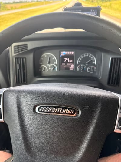A first-person perspective shot from inside a Freightliner truck shows the dashboard and steering wheel. The truck is traveling on a highway with a yellow field of crops on the left and a road stretching into the distance on the right. The dashboard features a digital display showing "71 mph" and other truck information, as well as analog gauges for speed and RPMs. A GPS device is mounted above the dashboard. The steering wheel is black and leather-wrapped, with the "FREIGHTLINER" logo prominently displayed in the center. The scene appears to be set in Williamstown, United States, based on the context provided, although the location is not explicitly visible in the image itself.