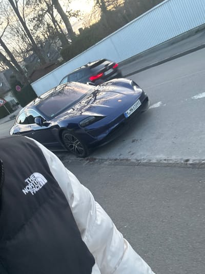 The scene depicts a street in Grünwald, Germany. A dark blue Porsche Taycan electric vehicle, bearing German license plate M MN 6189 E, is positioned at an angle on an asphalt road. Its front and left side are visible, with reflections of bare trees and a warm sky on its surface. In the background, a dark-colored SUV is observed driving on the same road, exhibiting slight motion blur. The foreground captures the upper torso and left arm of an individual, wearing a black and white jacket emblazoned with "THE NORTH FACE" logo on the left chest. The environment includes bare deciduous trees and residential structures in the background, alongside a light-colored, possibly corrugated, fence or wall. A circular traffic sign with a red border is partially visible on the left side of the road. The lighting indicates either early morning or late afternoon, with soft, directional light. The road surface consists of asphalt, bordered by a concrete curb.
