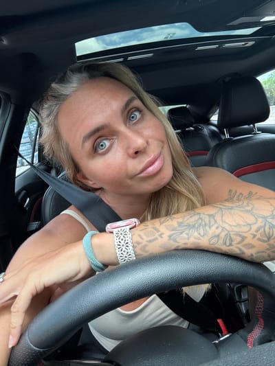 A woman with blonde hair and blue eyes is in the driver's seat of a car, looking at the camera. She is wearing a white tank top, a turquoise hair tie on her wrist, and a watch with a white floral band. Her left arm, which is tattooed, rests on the steering wheel. The car has black leather seats and a sunroof. The woman's seatbelt is visible. The image appears to be taken during the daytime, with natural light coming from the sunroof. The background is blurred but suggests an outdoor environment, possibly a street or parking lot. The location in Tha Chana, Thailand, is not discernible from the image itself.