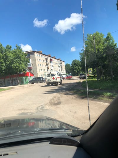 The image depicts a street scene in Izdeshkovo, Russia, on a clear, sunny day. In the foreground, the interior of a vehicle is visible, with the windshield reflecting buildings and trees. A tall, thin antenna extends upwards from the vehicle's exterior.

On the street, an older model white ambulance with a red cross emblem is parked. To the left of the ambulance, a dark-colored SUV is also parked, and a red sedan is seen further down the road. Across the street, a multi-story apartment building with brick detailing and numerous windows stands adjacent to a building with a red and green storefront, possibly a shop. Lush green trees line the street and the side of the road.

A yellow diamond-shaped road sign with the symbol of children walking is visible on the right side of the road, indicating a pedestrian crossing. The road surface is asphalt with some patchy wear. The sky is bright blue with a few scattered white clouds. Birds are visible as small dark specks in the sky.