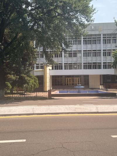 The image shows a building with a grid-like facade. A large tree partially obscures the left side of the building. To the right of the building is a road with a yellow curb. The photo appears to be taken in Kashgar mahalla, Uzbekistan. A decorative blue structure stands near the entrance of the building. A fence runs along the edge of the road.