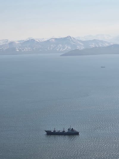 An aerial, high-angle shot captures a wide expanse of calm, gray-blue ocean under a hazy sky. In the distance, a range of snow-capped mountains forms a hazy, blue silhouette against the pale sky. In the mid-ground, a small ship is visible on the water. In the foreground, a larger, dark-colored cargo ship is positioned slightly to the right, its deck structures clearly defined. The water exhibits gentle ripples, catching the light in a way that suggests a bright, though overcast, day. The scene depicts a serene maritime landscape with a backdrop of formidable mountains, likely in a remote, coastal region of Russia, possibly near Крутобереговый based on the context provided.
