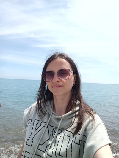 The image depicts an adult female positioned on a coastline in Krinitsa, Russia. She has dark brown, medium-length hair and wears large, purple-tinted sunglasses with metal side accents. A subtle smile is present on her face. A small dark mole is visible below her right eye and another on the right side of her neck/upper chest area. She wears a small gold hoop earring in her left ear. Her attire consists of a light grey-green, short-sleeved hooded sweatshirt (hoodie) featuring black distressed lettering, with visible partial characters including 'Y' and 'OG'. The hoodie's drawstring is visible. In the background, a calm blue-green sea extends to the horizon, meeting a light blue sky with sparse, wispy white clouds. A small, reddish-orange buoy is visible on the water in the distant left. The immediate foreground shows the edge of the water with gentle ripples and wet ground.