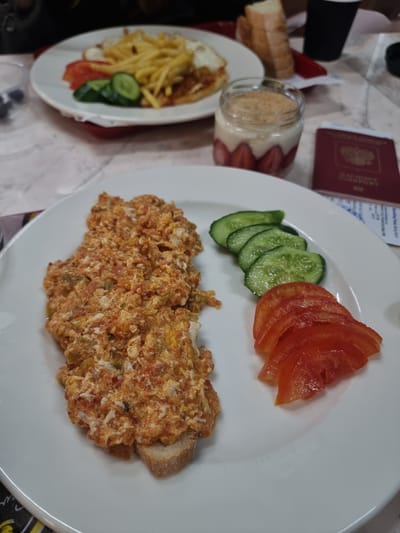 The image shows a table setting with food. There are two white plates, a jar, and a passport. The larger plate holds what appears to be scrambled eggs with tomatoes, sliced cucumbers, and tomatoes. The smaller plate has french fries, sliced cucumbers, and tomatoes with a fried egg. Beside the smaller plate is a jar of yogurt and strawberries, and a piece of bread. A passport is placed near the food. The environment appears to be a restaurant or dining area. There is no indication of the location being in Mora, Northern Cyprus/Cyprus.