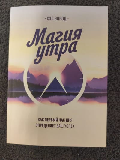 The image displays the cover of a book titled "Магия утра" (Magic of Morning) by Хэл Элрод (Hal Elrod). The book cover features a serene landscape with purple mountains reflecting in a calm body of water, with a stylized clock face overlaid. The text is in Russian. The book cover is placed on a dark gray, textured surface, likely a carpet or fabric. There are no people or any other objects visible in the image, and no information suggests its location in Schwäbisch Gmünd, Germany.
