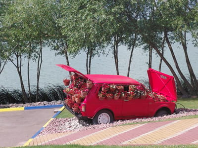 In Kovalivka, Ukraine, a vibrant red car has been transformed into a striking floral display. The car is parked outdoors, positioned on a bed of small, light-colored stones. Its rear hatch is open, revealing an abundance of potted flowers overflowing from within, extending to the edges of the opening. More potted flowers are affixed to the exterior of the car, adorning its sides and the front compartment where the hood is also lifted, showcasing yet more floral arrangements.

The car itself appears to be an older model, its once functional purpose now supplanted by its role as a decorative centerpiece. The flowers are a mix of pinks, whites, and reds, creating a striking contrast against the car's bold red exterior.

The car is situated in a landscaped area with a paved pathway composed of alternating yellow, pink, and beige rectangular tiles. This pathway runs diagonally across the foreground of the image. To the left of the car, a curb separates a paved area from the stone bed the car rests on. This curb is painted with bright yellow and blue stripes. Beyond the car, a body of water, likely a lake or river, stretches across the background, bordered by a dense line of tall, slender trees with abundant green foliage. The trees lean slightly, creating a dynamic visual line against the tranquil water. The sky above is a clear, pale blue, indicating fair weather. There are no people visible in the image.