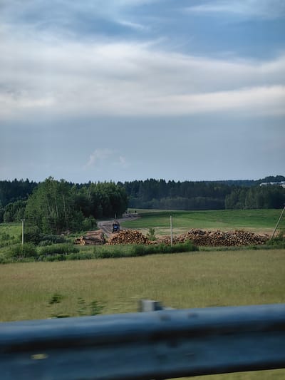 The image depicts a rural scene in the vicinity of Пятрышкаўскі сельскі Савет, Belarus. In the foreground, a blurred metal guardrail suggests the vantage point is from a moving vehicle. Beyond the guardrail, a grassy field transitions to an area with neatly stacked piles of felled logs, indicating logging activity. A blue forestry machine, possibly a harvester or forwarder, is positioned on a dirt track near the logs, with its mechanical arm extended. Several individuals are visible near the machinery and the log piles, engaged in the logging operation. A dense forest forms the backdrop, with a clear sky above dotted with clouds. The overall environment appears to be a mix of agricultural land and woodland, consistent with a rural Belarusian landscape.
