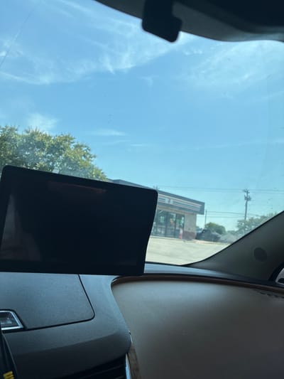 The image, taken from inside a vehicle in Balch Springs, United States of America, shows a view through the windshield. In the foreground, a black digital screen is mounted on the dashboard. Beyond the screen, a 7-Eleven convenience store is visible, characterized by its distinctive red, green, and white striped awning and signage. Trees and utility poles are also present in the background, under a partly cloudy blue sky. The interior of the car's dashboard, made of dark textured plastic and a brown leather-like material, is also visible.