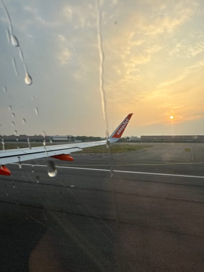 View from inside an airplane, possibly an EasyJet plane, on a runway at an airport, with rain drops visible on the window. The sun is setting, and clouds are visible in the sky. The location could not be determined in the city of Peschiera Borromeo.