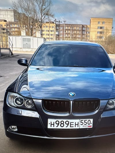 A dark blue BMW sedan is parked in front of apartment buildings in a city. The car's license plate reads "H989EH 550 RUS". The environment appears to be an urban setting with paved ground and a cloudy sky. The apartment buildings in the background are of various sizes and architectural styles, with some featuring yellow and beige facades. The image is taken from a slightly low angle, focusing on the front of the car. There are no people visible in the image. The scene appears to be a typical street view in a residential area.