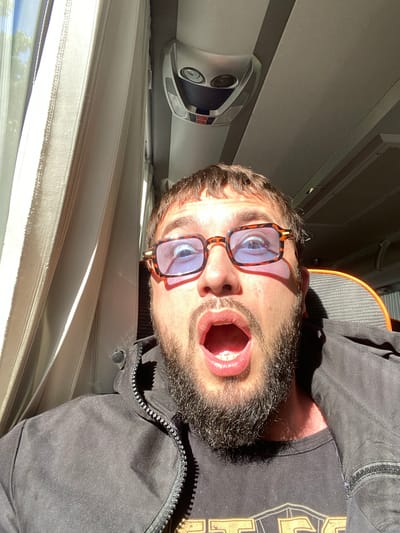 A man with a beard and glasses is taking a selfie inside what appears to be a bus. He is wearing a black jacket over a black t-shirt. His mouth is open in a surprised expression. The interior of the bus is visible, with a window with curtains on the left side, and overhead compartments and lighting fixtures on the ceiling. The man's eyes are wide open behind his blue-tinted glasses, and the lighting suggests it is daytime, possibly with sunlight coming through the window. The location is Seltso, Russia.