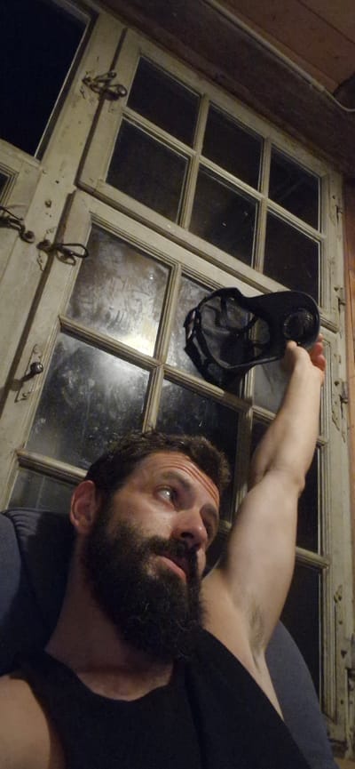 A man with a beard and a black tank top is sitting in a chair in front of a large, old, weathered window with multiple panes. He is holding a black mask in his raised right hand, with the mask partially obscuring the upper part of the window. The man is looking upwards and to his left. The window is set in a wooden frame, and there are metal latches on the left side of the window frame. The scene appears to be indoors, with dim lighting. The image does not provide specific information about a location in Wattenwil, Switzerland.