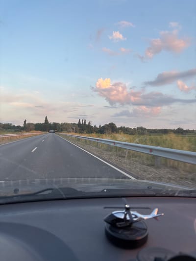This image depicts a view from inside a vehicle traveling on a highway. The foreground shows a portion of the dashboard with a decorative airplane model mounted on a suction cup. The majority of the image is a perspective shot of an empty asphalt road with white lane markings, stretching into the distance. On either side of the road are metallic guardrails and a grassy embankment dotted with trees. The sky above is a soft blue with scattered clouds tinged with pink and yellow, suggesting it is either sunrise or sunset. The lighting indicates it is likely late afternoon or early evening. The scene is calm and tranquil, with no other vehicles or people visible on the road. The overall impression is one of a peaceful journey through a rural or semi-rural landscape. No visible text is present in the image.