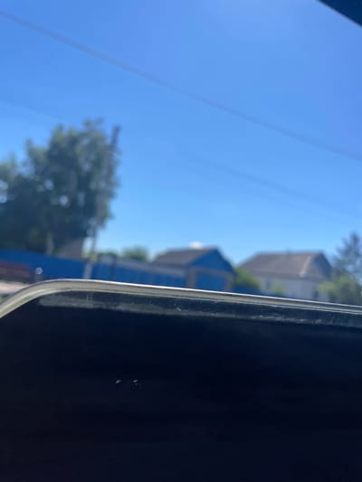 The image shows a blurry outdoor scene with a clear blue sky and a few trees in the background. In the foreground, there is a dark, reflective surface with a thin metallic stripe running across the top. The scene appears to be viewed through a window or from a car. The background includes the rooftops of several houses and a blue fence. The location is identified as Uspenka, Kazakhstan.