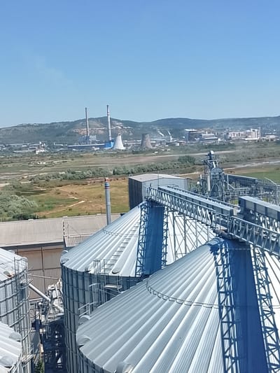The image shows a bright, clear day with a wide blue sky over a landscape dominated by industrial structures. In the foreground, several large, corrugated metal silos are visible, their conical roofs reflecting the sunlight. Metal walkways and support structures connect these silos. Behind the silos, a building with a metal roof is partially visible. Further in the background, a complex industrial facility stretches across the horizon. This facility includes tall smokestacks, cooling towers, and various interconnected buildings and infrastructure. The landscape surrounding the industrial area is characterized by rolling hills covered in greenery and fields of dry, light brown grass. The overall scene depicts an active industrial zone on a sunny day. The image is of the Maritsa East II thermal power plant and its associated facilities, located in the vicinity of Maritsa East mining complex, near the town of Radnevo, Bulgaria.