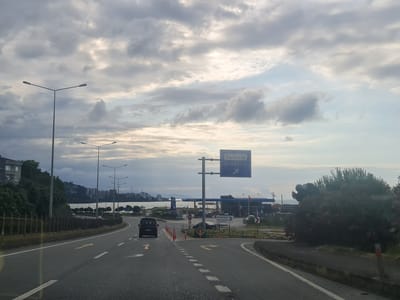 A cloudy sky looms over a coastal road in Araklı, Turkey. The scene is captured from the perspective of a vehicle moving along the asphalt. On the left side of the road, a multi-story building sits on a hillside covered in lush green trees, with the dark blue sea visible in the distance. Further along the road, streetlights punctuate the scenery, and a decorative metal fence lines the edge of the roadway.

A directional signpost stands prominently, displaying "Kalecik" and "Araklı" with an arrow pointing to the right. Adjacent to this sign is a gas station with pumps and a service building. Several vehicles are present, including a dark-colored van driving in the same direction as the viewer, and other cars parked or driving around the gas station. Traffic cones and barriers are strategically placed.

To the right, a large, dense green bush with pink flowers dominates the foreground, partially obscuring the view of the road and surrounding area. The weather appears overcast, with patches of brighter sky peeking through the clouds, suggesting it might be late afternoon or early morning. The overall mood is neutral, depicting a common roadside scene in a coastal town. The visible text includes place names on the signpost and potentially some branding on the gas station.