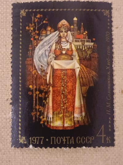 This is a postage stamp featuring a painted illustration of a woman in traditional Russian attire. She is adorned in a richly decorated red and gold dress, a white veil, and a jeweled headdress. The woman is holding a loaf of bread on a white cloth, presenting it forward. Behind her, a cityscape with onion-domed buildings, reminiscent of Moscow, is depicted, along with autumn foliage and what appear to be sheaves of wheat. The overall scene evokes a sense of tradition, abundance, and cultural heritage.

The stamp is bordered by a dark blue frame with a decorative yellow pattern along the edges. Visible text on the stamp includes "1977 • ПОЧТА СССР" (1977 • POSTAGE USSR) along the bottom edge, and vertically along the right side, "Н.М.Солоницина. Хлеб-соль. 1970" (N.M. Solonitsyna. Bread-salt. 1970). There is also a denomination of "4" followed by "к" (kopecks) in the lower right corner. The stamp itself appears to be printed on textured paper.