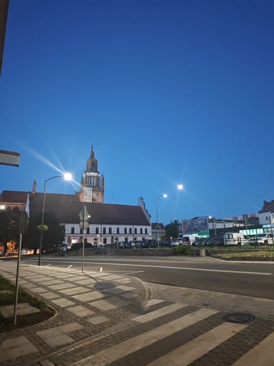 The image shows a street scene in Stargard, Poland, at dusk. In the background, a large, historic building with a tall tower dominates the skyline. The building appears to be a church or cathedral, with gothic architectural elements. In front of the building, there is a street with cars parked along the side. The street is paved with cobblestones and has a pedestrian crossing. The sky is a deep blue, suggesting it is either early morning or late evening. There are streetlights illuminating the scene, casting long shadows. The overall impression is one of a peaceful and historic urban environment.