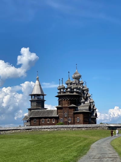 In Boyarshchina, Russia, a large wooden church with a bell tower stands behind a stone wall. The church features numerous domes and intricate wooden architecture. A paved path leads towards the church from the lower right of the frame. Two people are walking on the path, one in a brown robe and the other in white. The foreground is a lush green grassy field. The sky is bright blue with scattered white clouds.