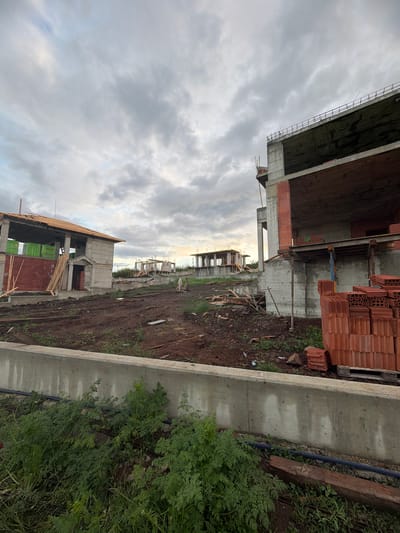 The image depicts a construction site in Cherni Vrah, Bulgaria, with unfinished buildings of reinforced concrete and brick. Stacks of bricks are visible near one of the structures. A concrete retaining wall runs along the edge of the site, separating it from an area with dense green vegetation. The ground is muddy and disturbed, typical of construction activity. Overcast skies fill the upper portion of the image.