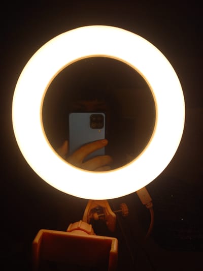In Sterlibashevo, Russia, a ring light illuminates a dark background. A smartphone, held by a hand, is visible in the reflection of the ring light's surface. The ring light is mounted on a light-colored clamp, suggesting it's a portable lighting accessory. The overall scene is dimly lit, with the bright ring light being the primary light source.