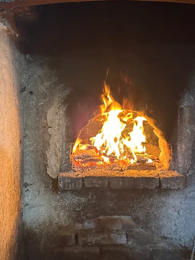 A fire is burning inside a masonry oven or fireplace. The opening to the fire is surrounded by rough-textured stone or brickwork. The flames are bright yellow and orange, filling the opening. The background is dark, suggesting the interior of the oven is dimly lit. The location is unknown and could not be determined.