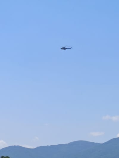 A blue helicopter is captured in flight against a clear, light blue sky. The helicopter is positioned in the upper middle section of the frame, appearing as a small dark silhouette. It has a rotor on top and a tail rotor. The scene is dominated by the vast expanse of the sky, with only the very top of a mountain range visible along the bottom edge of the image. The mountains are a muted blue-grey, suggesting they are some distance away. There are a few wispy white clouds scattered in the sky, particularly in the lower half of the frame, contrasting with the otherwise uniform blue. The overall impression is of a peaceful and open sky. The time of day appears to be daytime, likely mid-morning or afternoon, given the bright, clear sky. There is no indication of weather conditions other than clear skies. No people are visible in the frame, and no text is present. The location is identified as Kurtovo Konare, Bulgaria, but this context is not directly visible in the image itself.