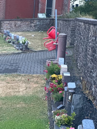 The image depicts a cemetery in Wadern, Germany. In the foreground, a row of gravestones with flowers and urns is visible. To the left of the gravestones is a paved walkway and a grassy area. In the background, two red watering cans are hanging on a metal pole. A stone wall runs along the back of the cemetery, with a building made of red brick behind it. The overall scene suggests a peaceful and well-maintained burial ground.