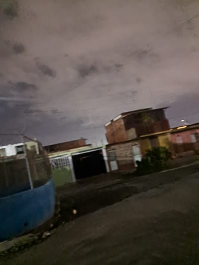 The image depicts an outdoor scene at night in what appears to be a residential area, possibly within the city of Cumana, Venezuela, based on the context provided. The sky is overcast with dark clouds, and there are faint lines suggesting power lines. In the foreground, a dark, textured surface, likely a road or unpaved ground, dominates the lower portion of the image. To the left, a large, curved structure painted in a vibrant blue color is visible, its texture suggesting it might be a water tank or a similar large object. Behind the blue structure, a building with a white facade and darker window openings is partially visible. Further into the background, several structures with brick exteriors and metal roofing are seen. Some of these buildings have visible windows and doors, and one appears to be a multi-story dwelling. There is also a hint of greenery, possibly bushes or small trees, clustered around the base of one of the buildings. The overall lighting suggests a low-light environment, typical of nighttime, with artificial lights likely illuminating some of the structures. No people are visible in the image. The scene conveys a sense of urban or semi-urban residential environment under a cloudy night sky.