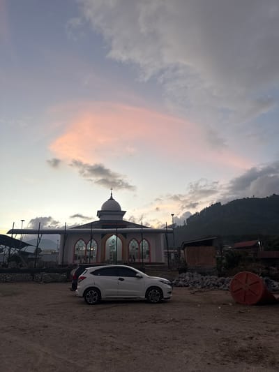 In the city of Mendale, Indonesia, a mosque stands prominently against a twilight sky. The mosque features a domed roof, arched windows with intricate detailing, and a light-colored facade. In the foreground, a white car is parked on a dirt lot adjacent to the mosque. The lot is cluttered with piles of rocks and a large orange cylindrical object. A gently sloping, tree-covered hillside rises behind the mosque and surrounding buildings. The sky displays soft hues of pink and orange, characteristic of sunset, with scattered clouds.