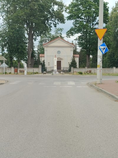 The image shows a street scene in Łomna, Poland. In the background, a small, cream-colored building with a red-tiled roof stands behind a low stone wall. The building has a central arched entrance and a circular window above it. A statue of a robed figure stands to the right of the entrance. Two large trees with dense green foliage frame the building on either side.

In the foreground, a paved road with a pedestrian crossing is visible. A light-colored utility pole is situated on the right side of the road, displaying two traffic signs: a yellow triangle pointing downwards and a blue square with a white silhouette of a person walking. The area behind the wall appears to be a grassy verge with some bushes and two wooden benches. The sky above is overcast.
