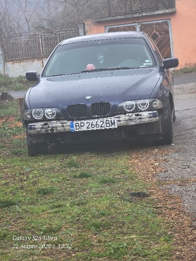 A dark blue BMW E39 sedan is stationary on a dirt and gravel surface in Dolna Beshovitsa, Bulgaria. The vehicle faces the observer, displaying a damaged front bumper with exposed undercarriage material and modified headlights featuring "angel eyes." A Bulgarian license plate, "BP 2662 BM," is mounted on the front. Water droplets are visible on the hood and windshield. A pink object and a hanging ornament are discernible on the dashboard through the windshield. The immediate foreground consists of patchy green grass and exposed soil. In the background, to the left, a rusted metal fence or gate is visible, with bare trees beyond it under an overcast sky. To the right, an orange-colored building with a weathered, double-leaf wooden garage door featuring a diamond pattern is present. No individuals are depicted.