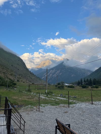 A scenic view of the Tegenekli Gorge in Russia is depicted. The foreground features a gravel path leading to a wooden bench and a metal staircase. The midground reveals a grassy field with scattered rocks and a simple fence structure. Beyond the field, power lines stretch across the landscape, supported by wooden poles. In the distance, majestic mountains dominate the scene, their peaks partially obscured by clouds. The sky is a vibrant blue, dotted with white, fluffy clouds, some of which are illuminated by sunlight, casting a warm glow on the mountain slopes. The lush green vegetation on the mountainside suggests a temperate climate.