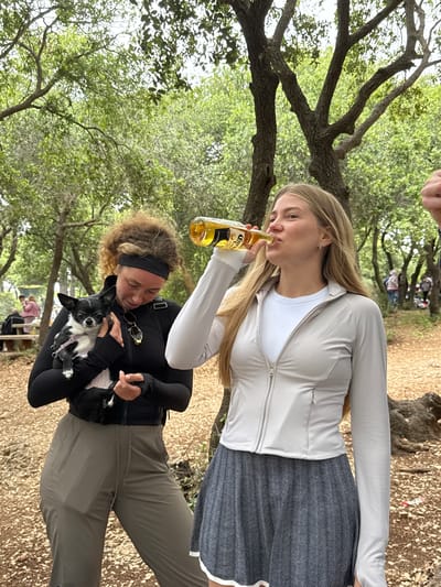 The photograph, taken outdoors in a wooded area, depicts two women. The woman in the upper portion of the frame is blonde and wearing a light-colored zip-up jacket, a white undershirt, and a dark herringbone skirt. She is drinking from a bottle of Corona beer. The woman in the lower portion of the frame is wearing a black long-sleeved shirt, dark pants and a black headband. She is holding a small black and white dog. The surroundings are characterized by trees with green foliage and a ground covered in leaves. Other individuals are indistinctly visible in the background. Although asked to include information from Nesher, Israel, there is no identifiable information in the image to locate the scene in that city.