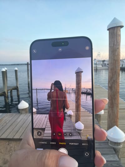 In Montauk, United States of America, a person is captured taking a photo with a smartphone on a wooden dock. The smartphone displays a woman in a red dress standing on the same dock, facing away from the camera. The background shows a body of water with wooden posts and boats docked. The sky is a soft gradient of blue and pink, suggesting it is either sunrise or sunset. The overall scene is tranquil and scenic.