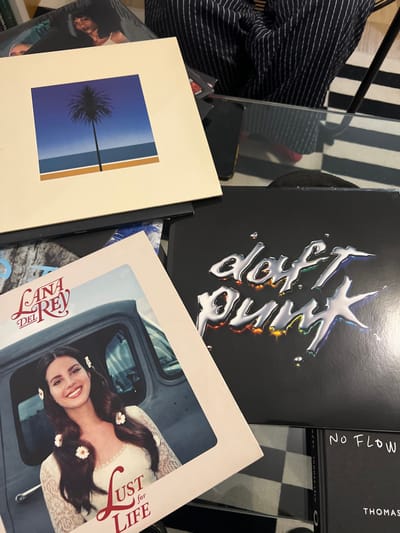 The image shows several album covers stacked on top of each other. Lana Del Rey's "Lust for Life" is visible, along with Tinashe's "Songs For You" and "Endless Summer" by Alan Walker. A book with the title "NO FLOW" is stacked on the side. The background includes a striped cushion and a checkered floor. A cell phone is also seen in the background.
