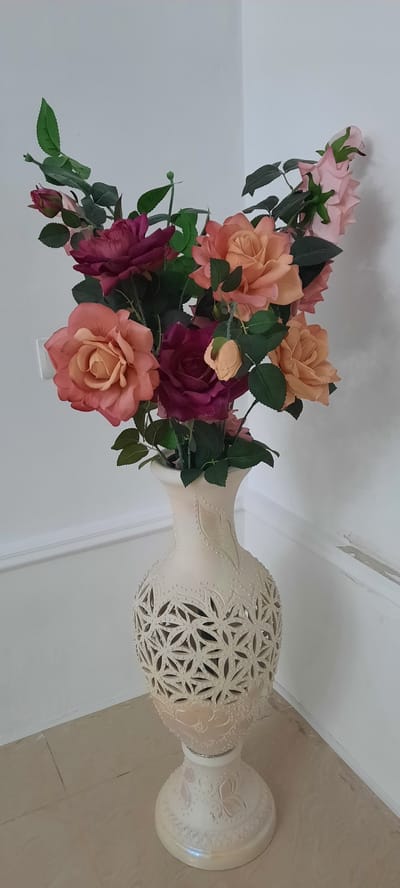 A vase filled with artificial roses stands on a tiled floor, leaning against a white wall. The vase is a light, creamy color and has an ornate, cut-out pattern around its body, resembling latticework. There are also embossed floral designs on the base of the vase. The roses are a mix of colors, including pink, peach, and deep pink, with green leaves. The scene appears to be indoors, likely in a home or commercial setting. The lighting suggests it could be daytime, but there are no specific weather or time-of-day cues. The location is identified as Gudermes, Russia. No people are visible in the image, and there is no visible text.