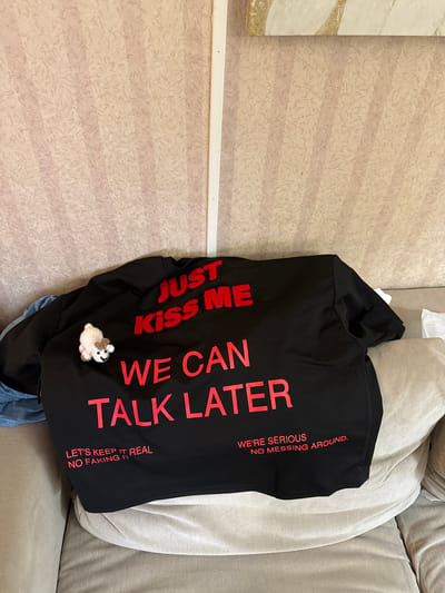 The image shows a black t-shirt with red lettering draped over a couch. The t-shirt has text that reads "JUST KISS ME WE CAN TALK LATER" in large letters. Below this, in smaller letters, it says "LET'S KEEP IT REAL NO FAKING IT" on the left side and "WE'RE SERIOUS NO MESSING AROUND." on the right side. A small crocheted white animal figurine is pinned to the left breast area of the t-shirt. The background is a light-colored textured wallpaper. The image is taken from a slightly elevated angle, looking down at the t-shirt on the couch. There is no information in the image to suggest the location is Hampton, United States of America.