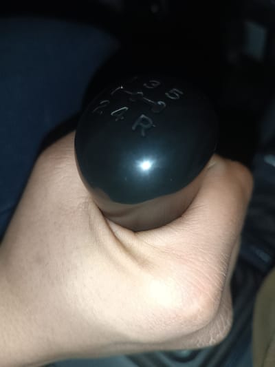 The image displays a close-up view of a human hand gripping a black gear shift knob, likely in the interior of a vehicle. The hand, which appears to be light-skinned, encircles the base of the knob with fingers visible on the left side and the thumb on the right. The skin shows natural texture and folds, particularly around the knuckles and where the hand meets the gear knob.
The gear shift knob itself is smooth, black, and has a noticeable reflective glare on its surface, indicating a light source directed towards it. Imprinted on the top surface of the knob is a standard five-speed manual transmission shift pattern. The pattern clearly shows the numbers "1," "3," and "5" arranged in the upper row, and "2," "4," and "R" (for Reverse) in the lower row. Lines connect these positions, illustrating the shifting gates. A central vertical line connects the first and second gears, third and fourth gears, and fifth and reverse gears. Horizontal lines connect 1-3, 3-5, 2-4, and 4-R.
The background of the image is largely dark and out of focus, suggesting a low-light environment, possibly the interior of a car at night or in a dimly lit space. To the left of the hand, a blurred dark blue or black fabric texture can be seen, which might be clothing or part of the car's interior. The overall impression is that someone is actively engaging with the car's manual transmission.
